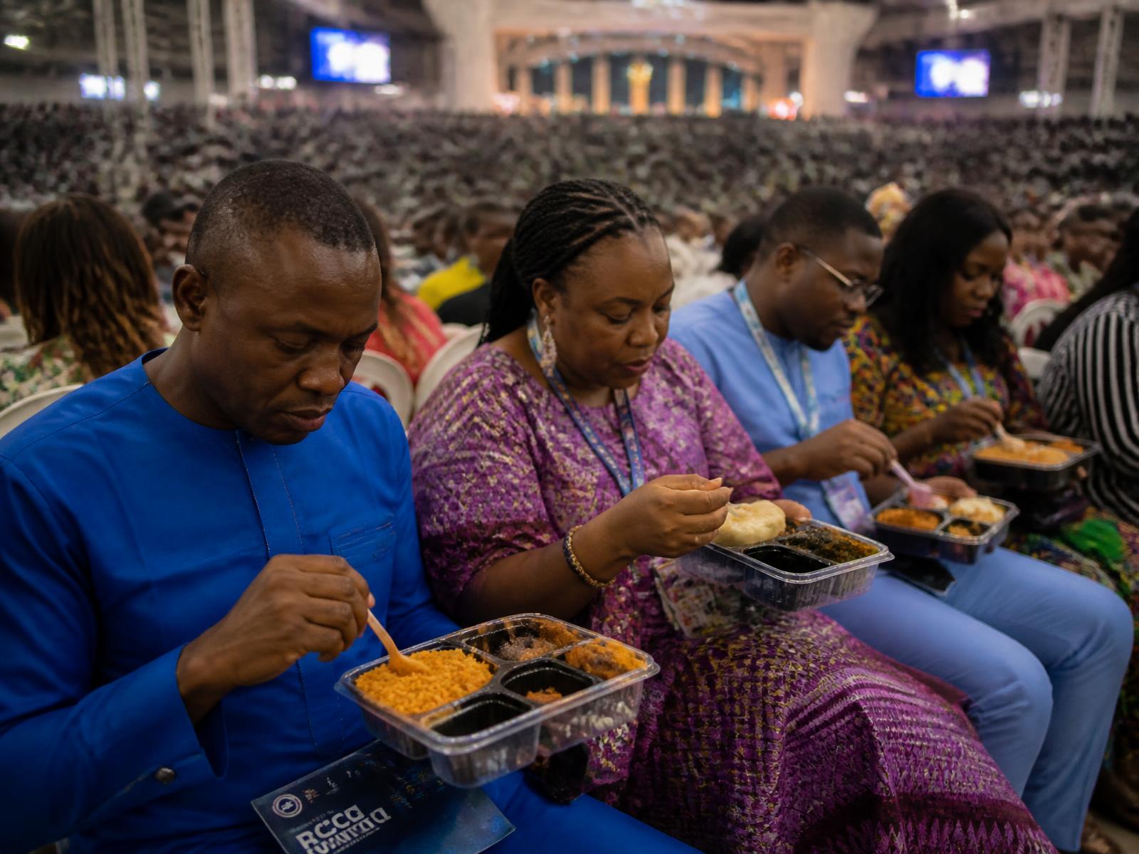 A close-up of RCCG convention attendees seated in the packed Redemption City auditorium, eating rich Nigerian meals from unbranded takeaway containers while facing the distant altar.